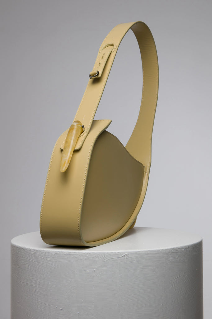 Flat topped oval structured saddle bag with bio resin accessories and non-removable but adjustable calf leather strap on a white stand