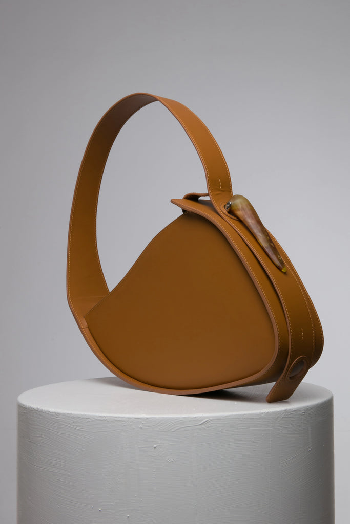 Flat topped oval structured saddle bag with bio resin accessories and non-removable but adjustable calf leather strap on a white stand