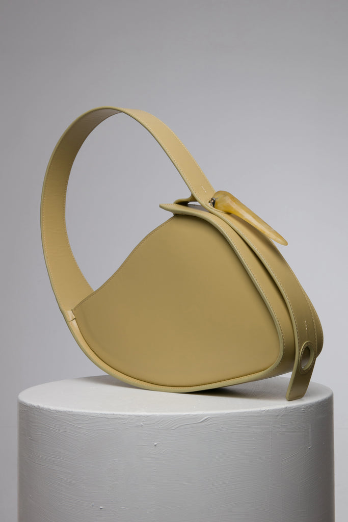 Flat topped oval structured saddle bag with bio resin accessories and non-removable but adjustable calf leather strap on a white stand