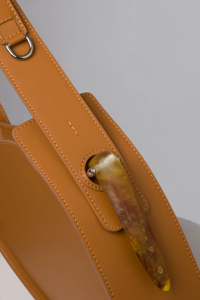 Flat topped oval structured saddle bag with bio resin accessories and non-removable but adjustable calf leather strap on a white stand